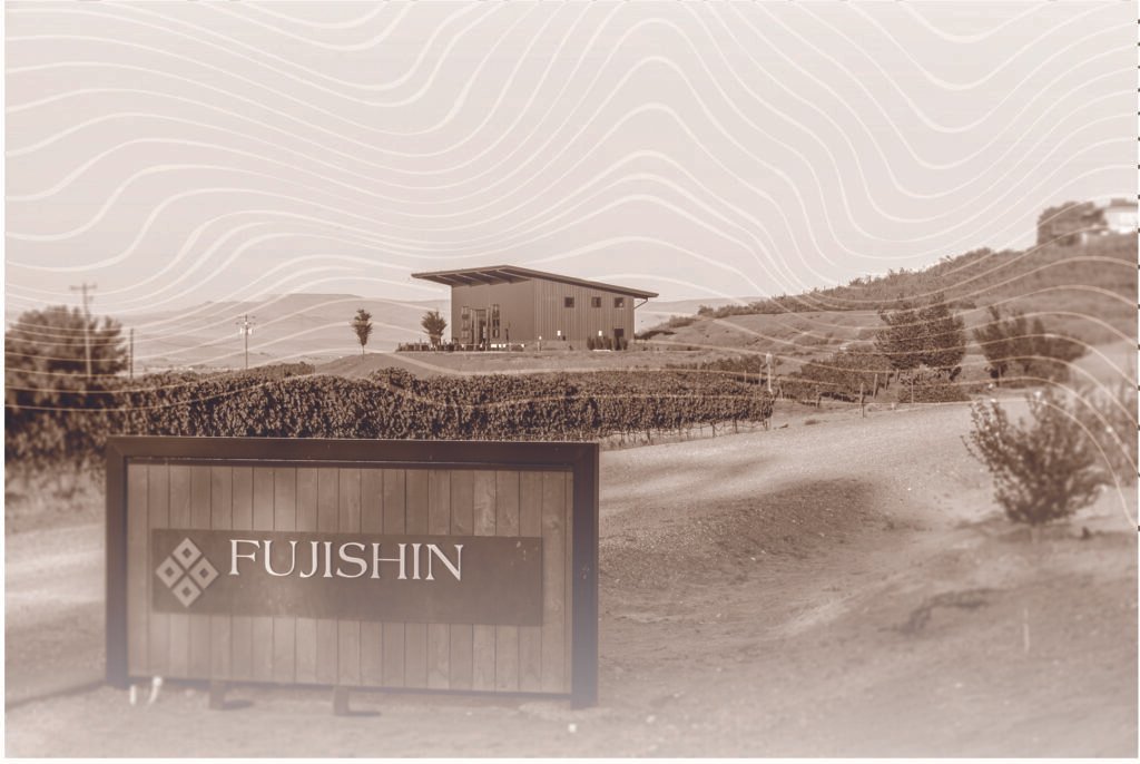 A wooden sign with the word "Fujishin" in the foreground with vineyard and building behind it